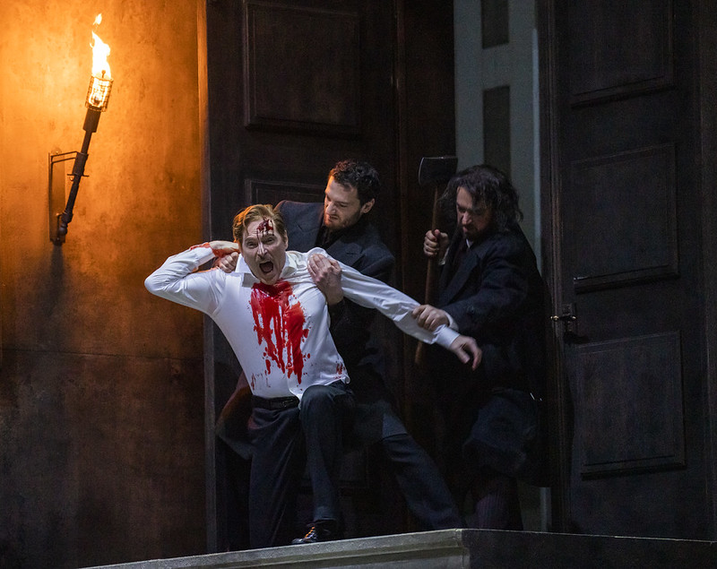 A man in a white shirt stained with blood is held by two other men in dark clothing near a door, one raising an axe, against a dimly lit background with a torch.