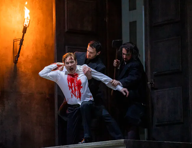 A man in a white shirt stained with blood is held by two other men in dark clothing near a door, one raising an axe, against a dimly lit background with a torch.