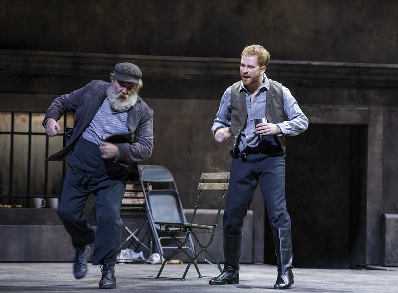 Two men dance on stage. One man stands on the right and is wearing navy trousers and a waistcoat. An older man is demonstrating a dance move. He is wearing a dark grey suit jacket and navy trousers. He has a full white beard. 