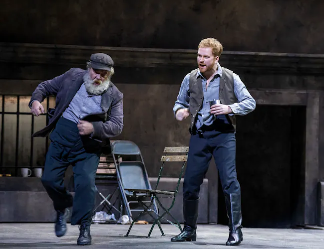 Two men dance on stage. One man stands on the right and is wearing navy trousers and a waistcoat. An older man is demonstrating a dance move. He is wearing a dark grey suit jacket and navy trousers. He has a full white beard.