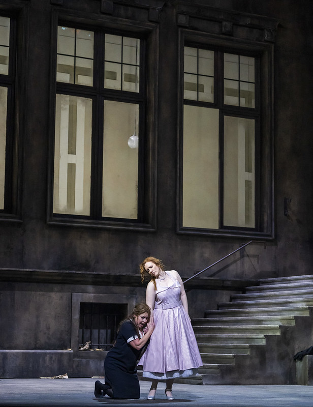 Elektra, in a black maid's dress kneels at the feet of a woman in a tea length purple dress. Elektra is gripping the womans hand. There is a dark stone wall behind them. 