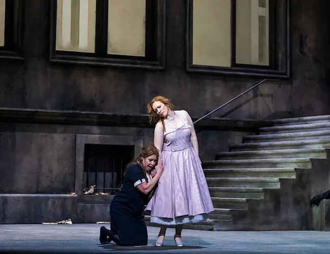 Elektra, in a black maid's dress kneels at the feet of a woman in a tea length purple dress. Elektra is gripping the womans hand. There is a dark stone wall behind them.