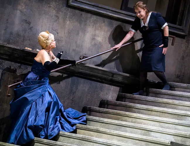 Two women interact on a staircase; one wears a formal electric blue gown and black silk gloves, the other stands above her in a black maid's dress.