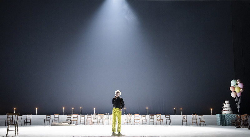 A clown in lime green trousers and a black shirt stands in a spotlight onstage. There are empty wooden chairs lining the back of the stage. 