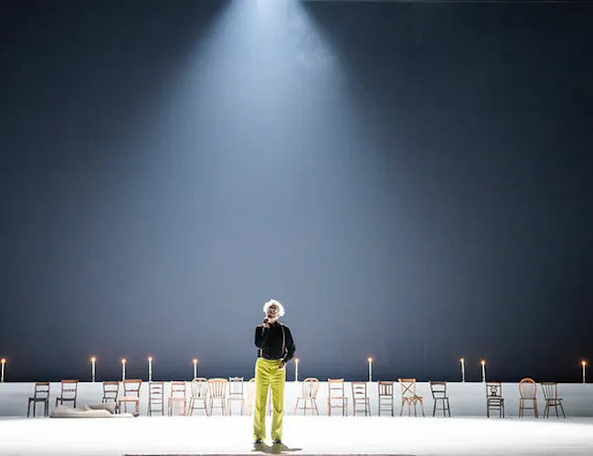 A clown in lime green trousers and a black shirt stands in a spotlight onstage. There are empty wooden chairs lining the back of the stage.