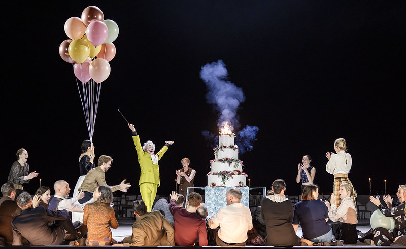 A clown in a lime green suit presents a four tier cake to a group of onlookers who clap as the top tier is lit with a sparkler. 
