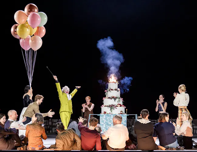 A clown in a lime green suit presents a four tier cake to a group of onlookers who clap as the top tier is lit with a sparkler.