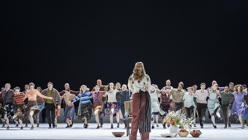 A line of people have their arms over each shoulders as they run towards the front of the stage. A woman wearing brown trousers and a floral top stands with her back to the camera as she waits for them to arrive. 