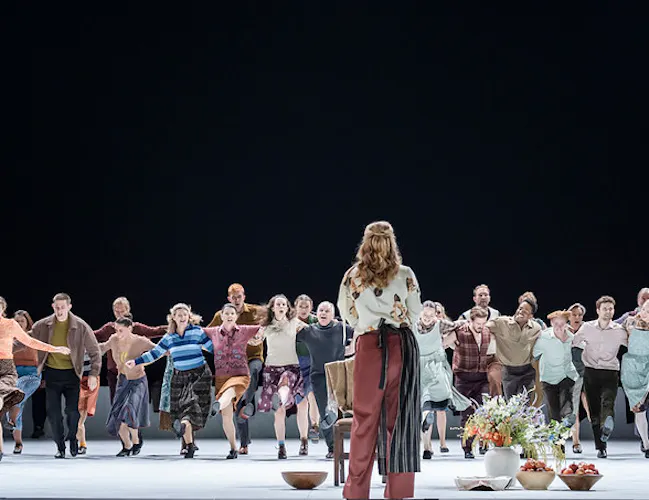 A line of people have their arms over each shoulders as they run towards the front of the stage. A woman wearing brown trousers and a floral top stands with her back to the camera as she waits for them to arrive.