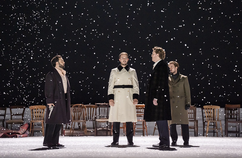 Four men stand onstage as snow falls around them. Two men stand facing each other with guns in their hands pointing down, ready for a duel. One man in a light cream coat stands centre stage, postioned between the two of them as a referee. 