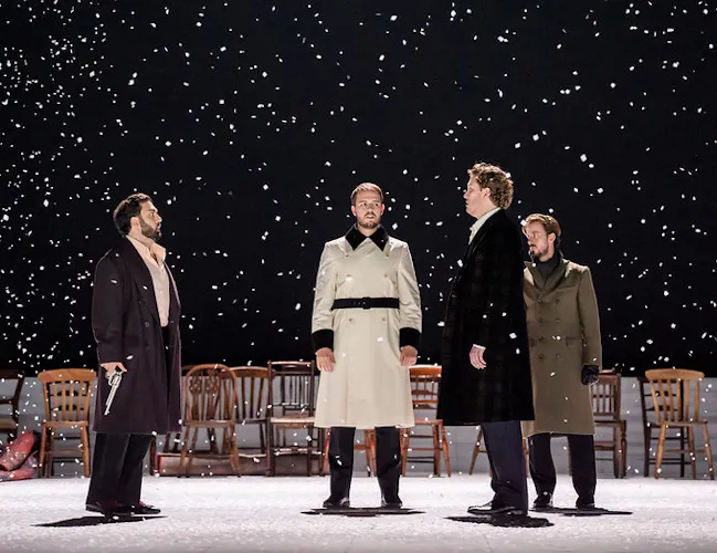 Four men stand onstage as snow falls around them. Two men stand facing each other with guns in their hands pointing down, ready for a duel. One man in a light cream coat stands centre stage, postioned between the two of them as a referee.