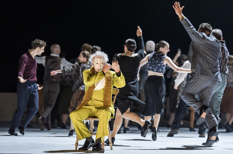A clown in a lime green suit is putting on white face paint as a large group of people dance behind him 