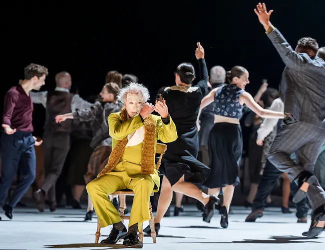 A clown in a lime green suit is putting on white face paint as a large group of people dance behind him