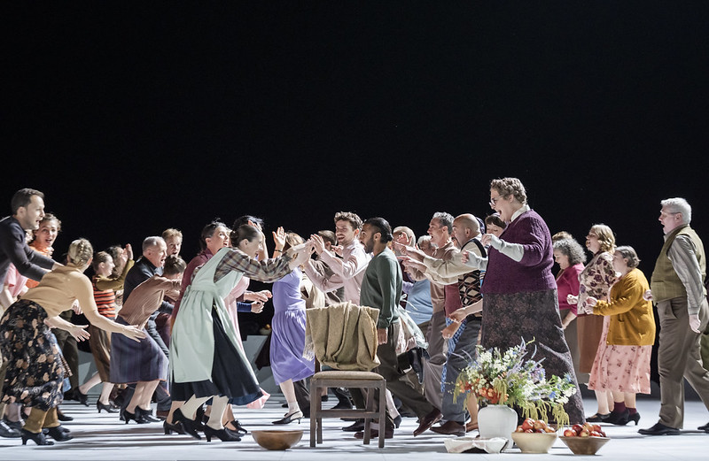 Lots of people dance on stage. There is an empty chair with a green jacket over it centrestage. 