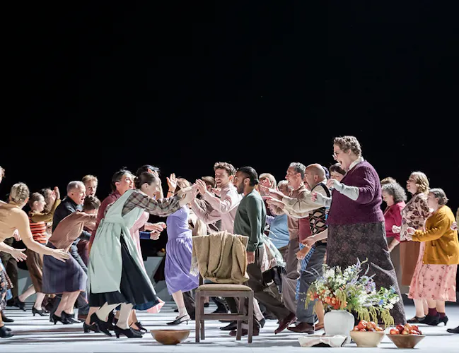 Lots of people dance on stage. There is an empty chair with a green jacket over it centrestage.
