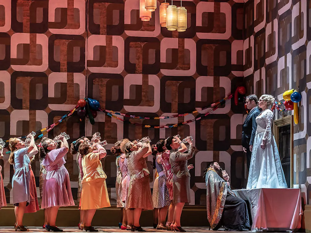 A group of people in party clothes take a drink in unison. They are toasting the married couple who are standing on a table. There are paper chains on the wall.