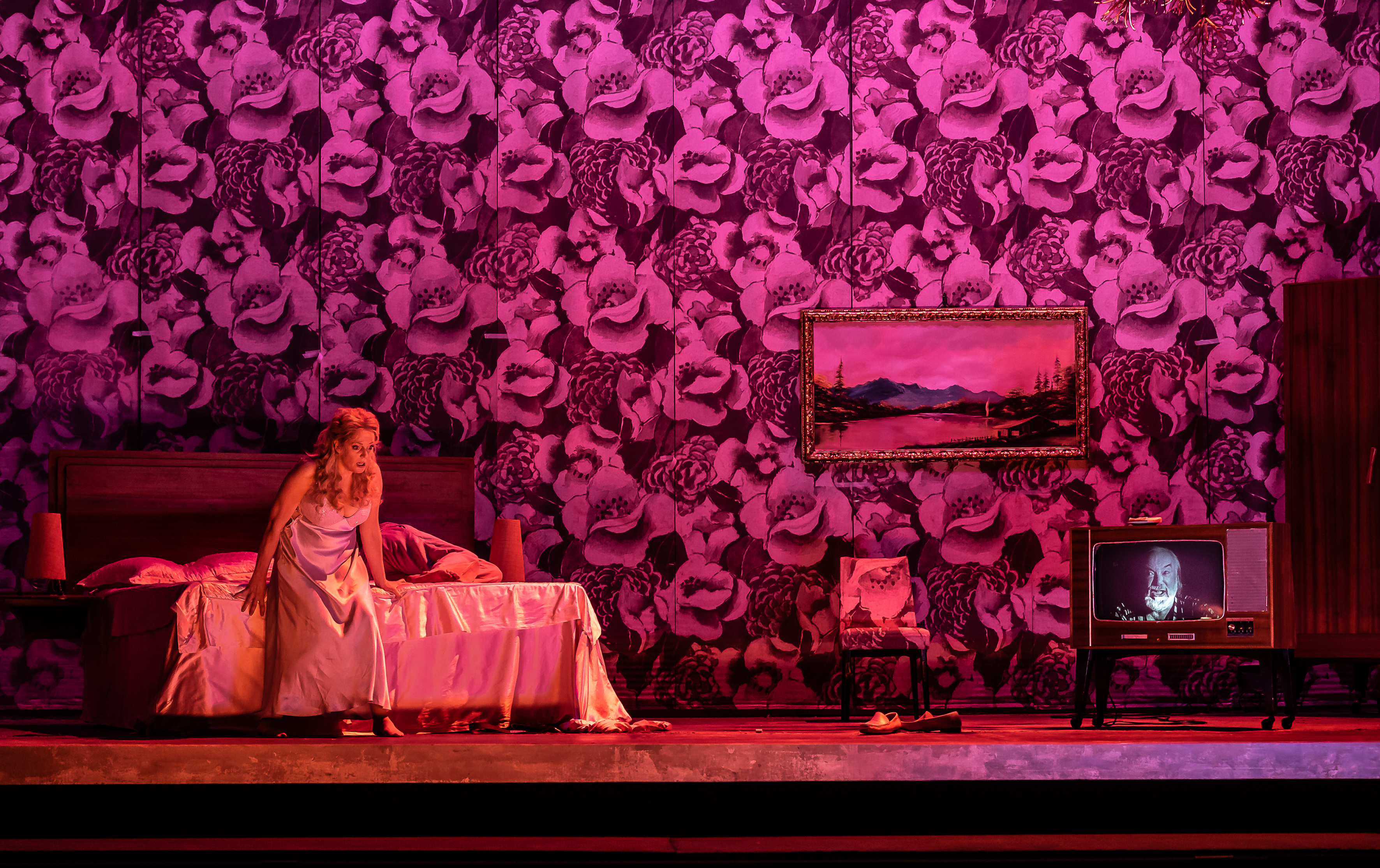 A woman sits on the end of the bed in a white nightgown. The room is pink. There is a man's face on the television. 