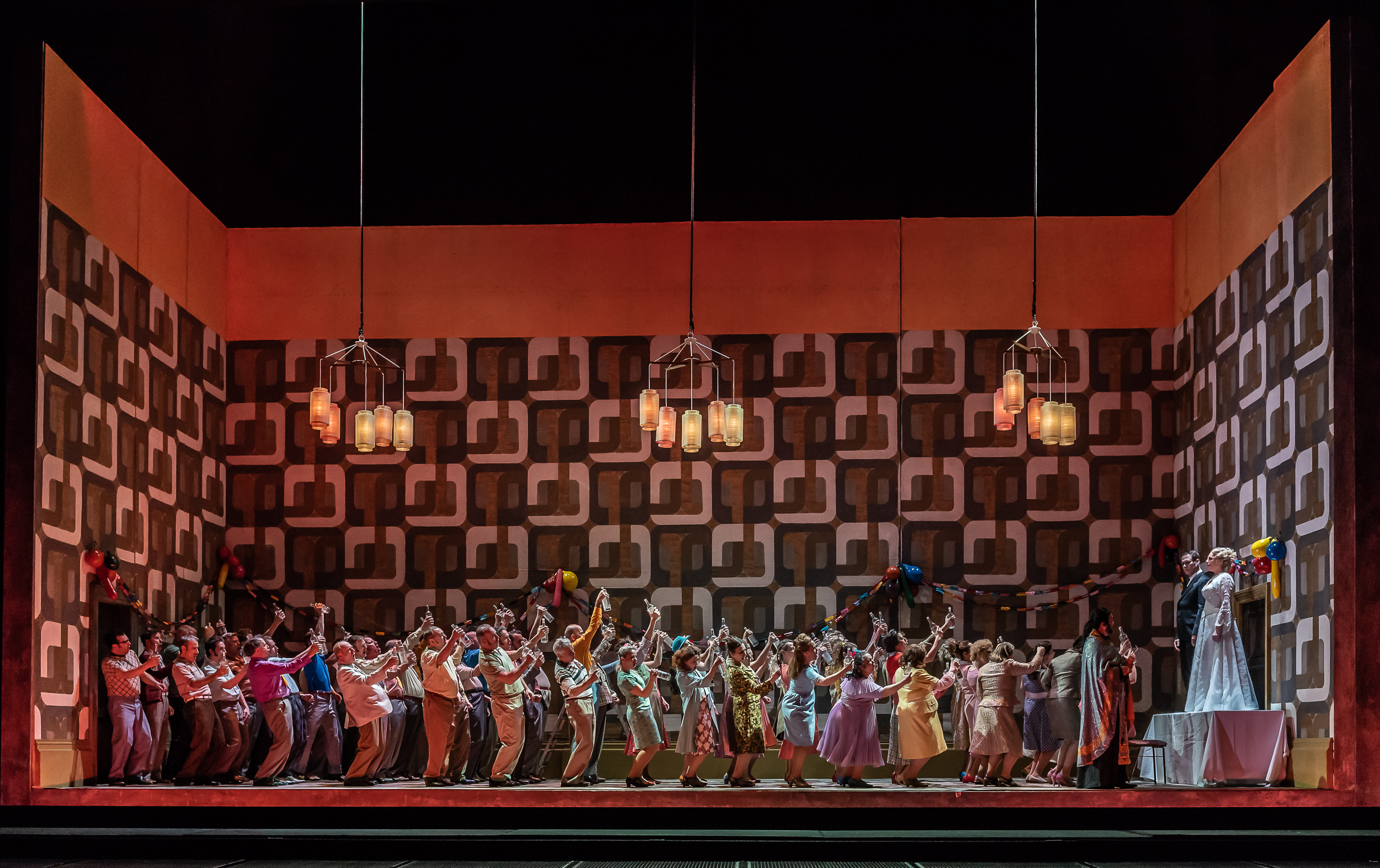 A group of people raise their glasses. They are toasting the married couple who are standing on a table. There are paper chains on the wall. 