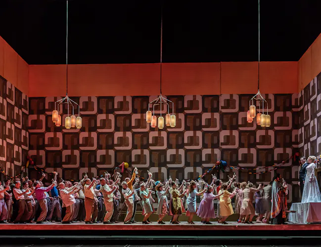 A group of people raise their glasses. They are toasting the married couple who are standing on a table. There are paper chains on the wall.