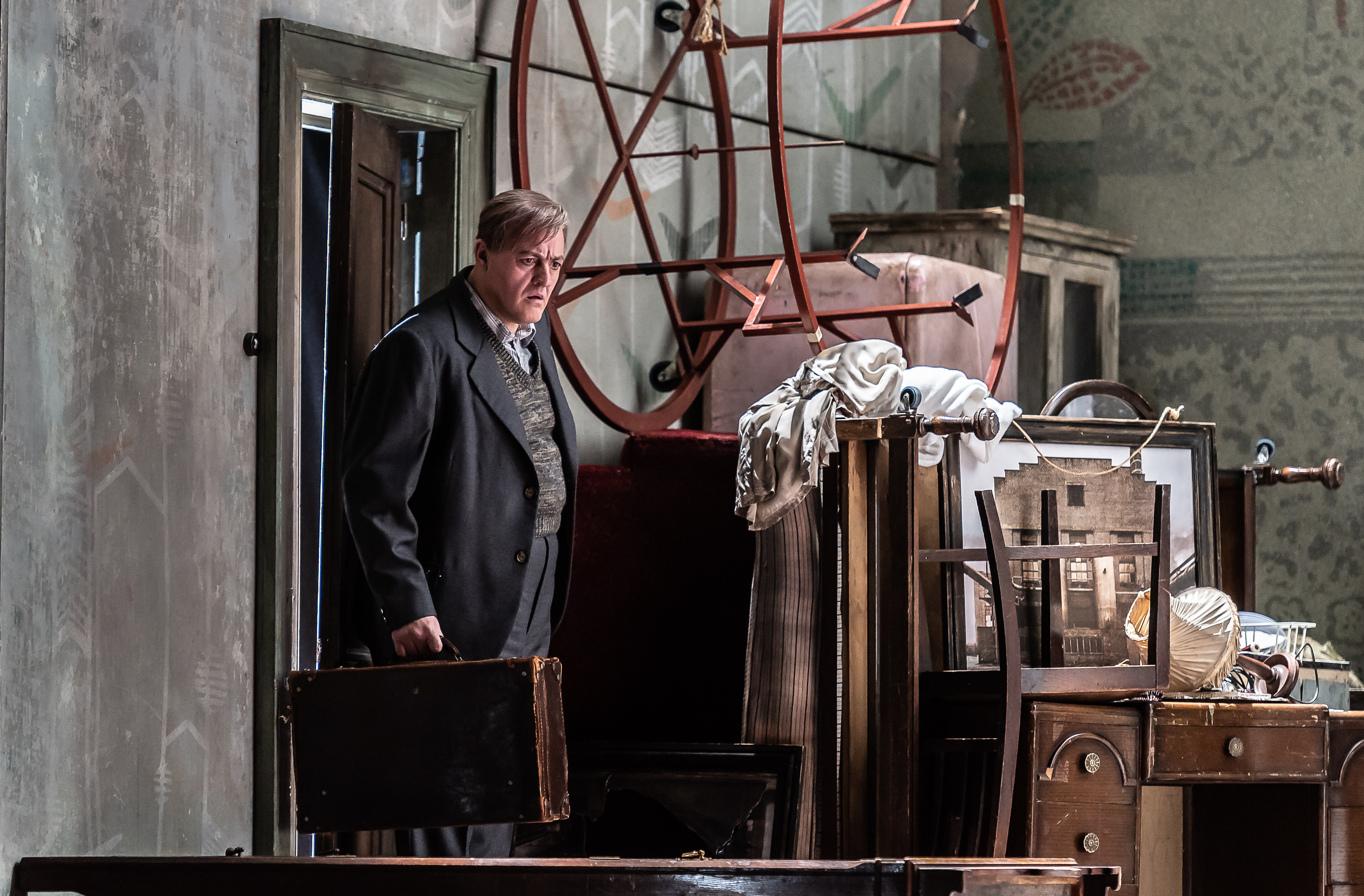 A man wearing a grey suit steps into a storage room. He carries a briefcase. The room has lots of boxes and frames covered in sheets. 