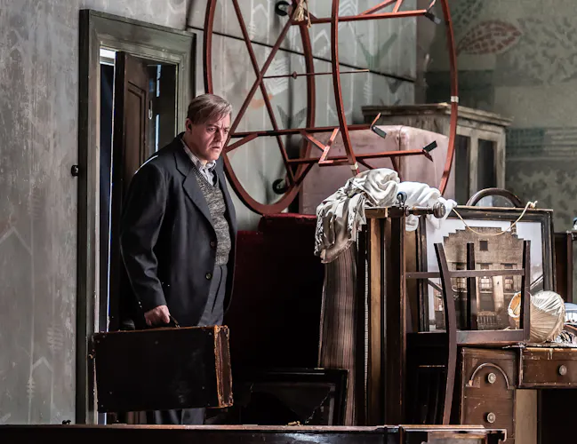 A man wearing a grey suit steps into a storage room. He carries a briefcase. The room has lots of boxes and frames covered in sheets.