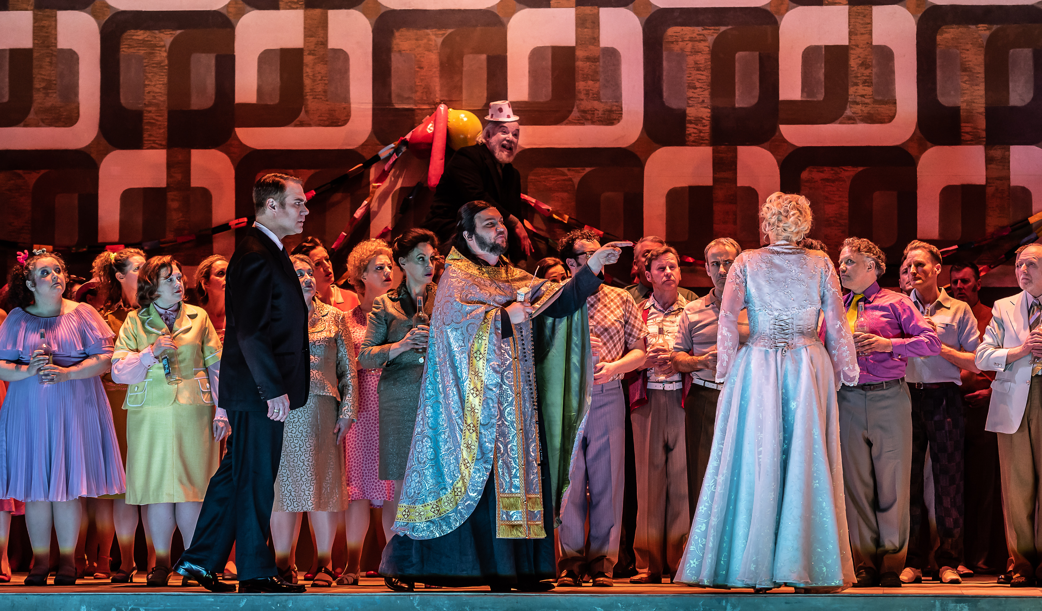 A man in priests robes gestures towards a woman in a white dress who stand with her back to the audience. A man in a black suit is standing watching her. 
