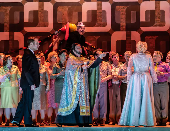 A man in priests robes gestures towards a woman in a white dress who stand with her back to the audience. A man in a black suit is standing watching her.
