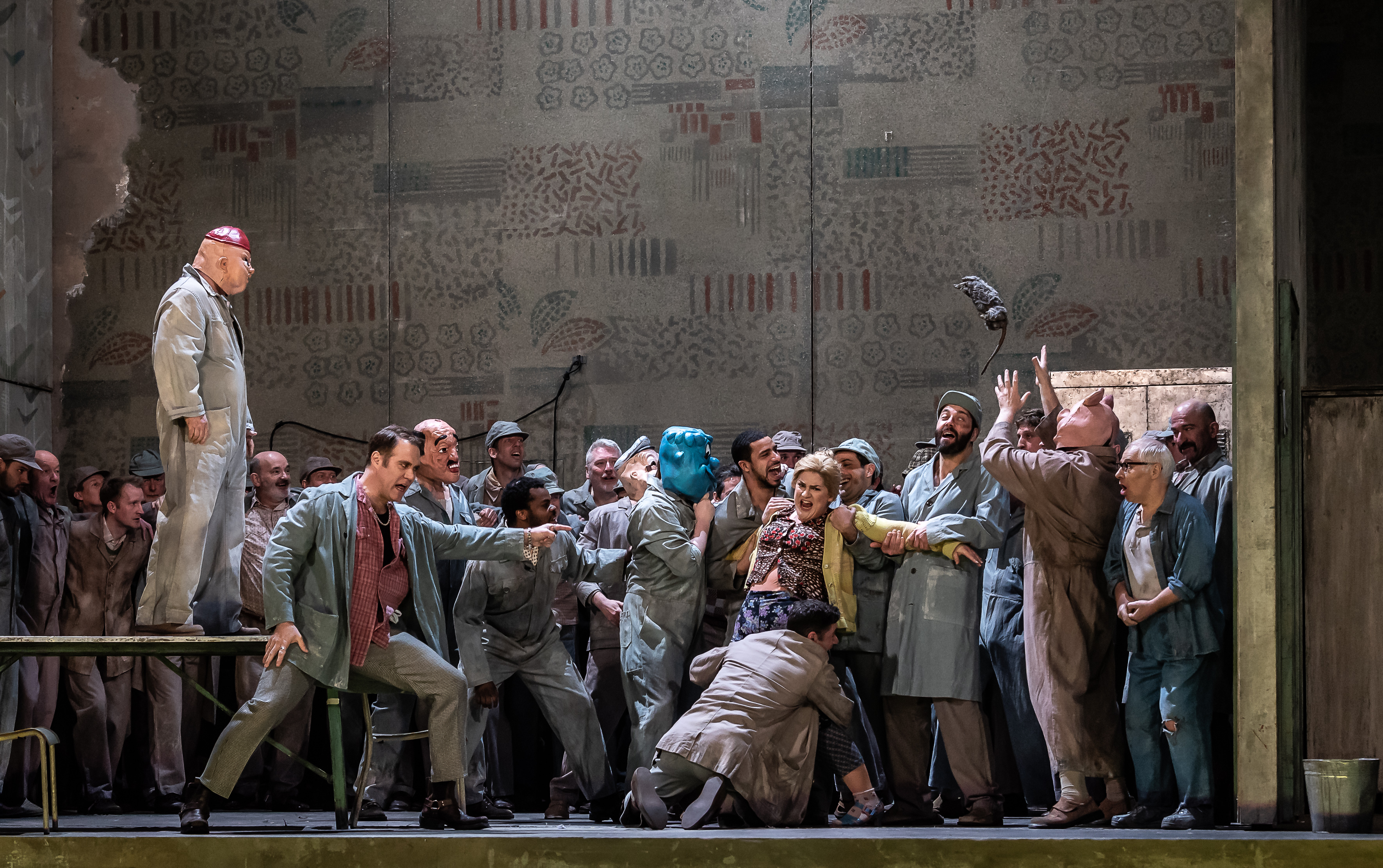 A group of men in grey boiler suits and animal masks restrain a woman wearing a yellow coat. A man wearing a pig mask stands on a table. Another man throws a rat into the air. 