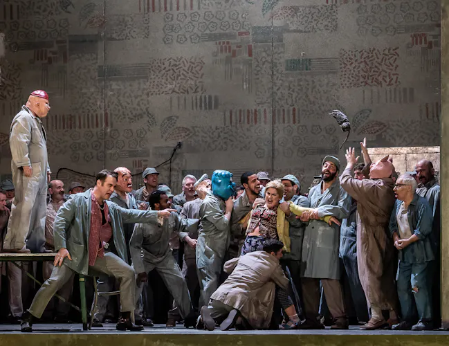A group of men in grey boiler suits and animal masks restrain a woman wearing a yellow coat. A man wearing a pig mask stands on a table. Another man throws a rat into the air.