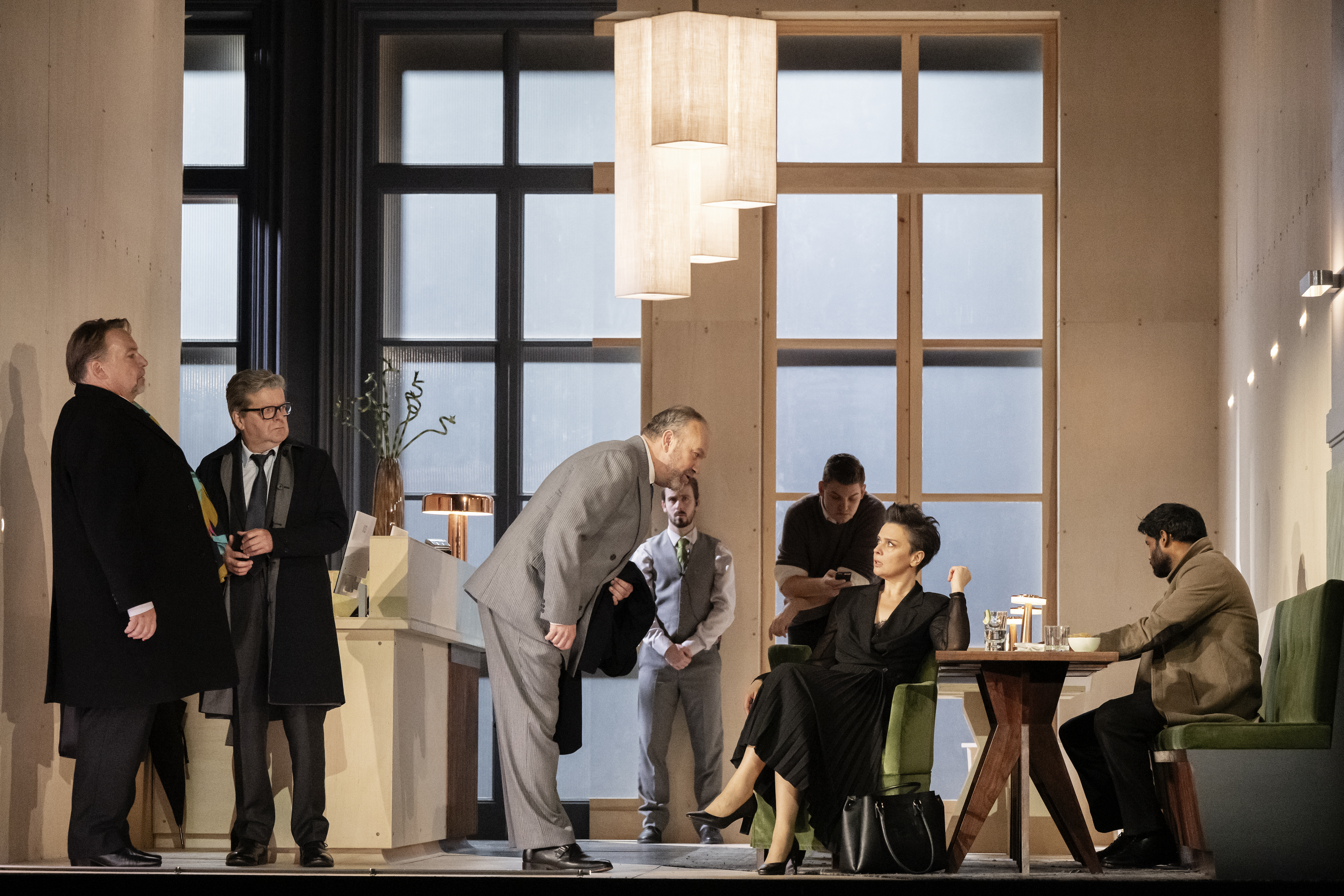 A group of people sit in a modern and bright room. A man in a grey suit leans down to speak to a woman in a black dress who is sat at a table. 