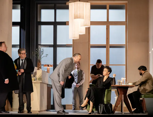 A group of people sit in a modern and bright room. A man in a grey suit leans down to speak to a woman in a black dress who is sat at a table.