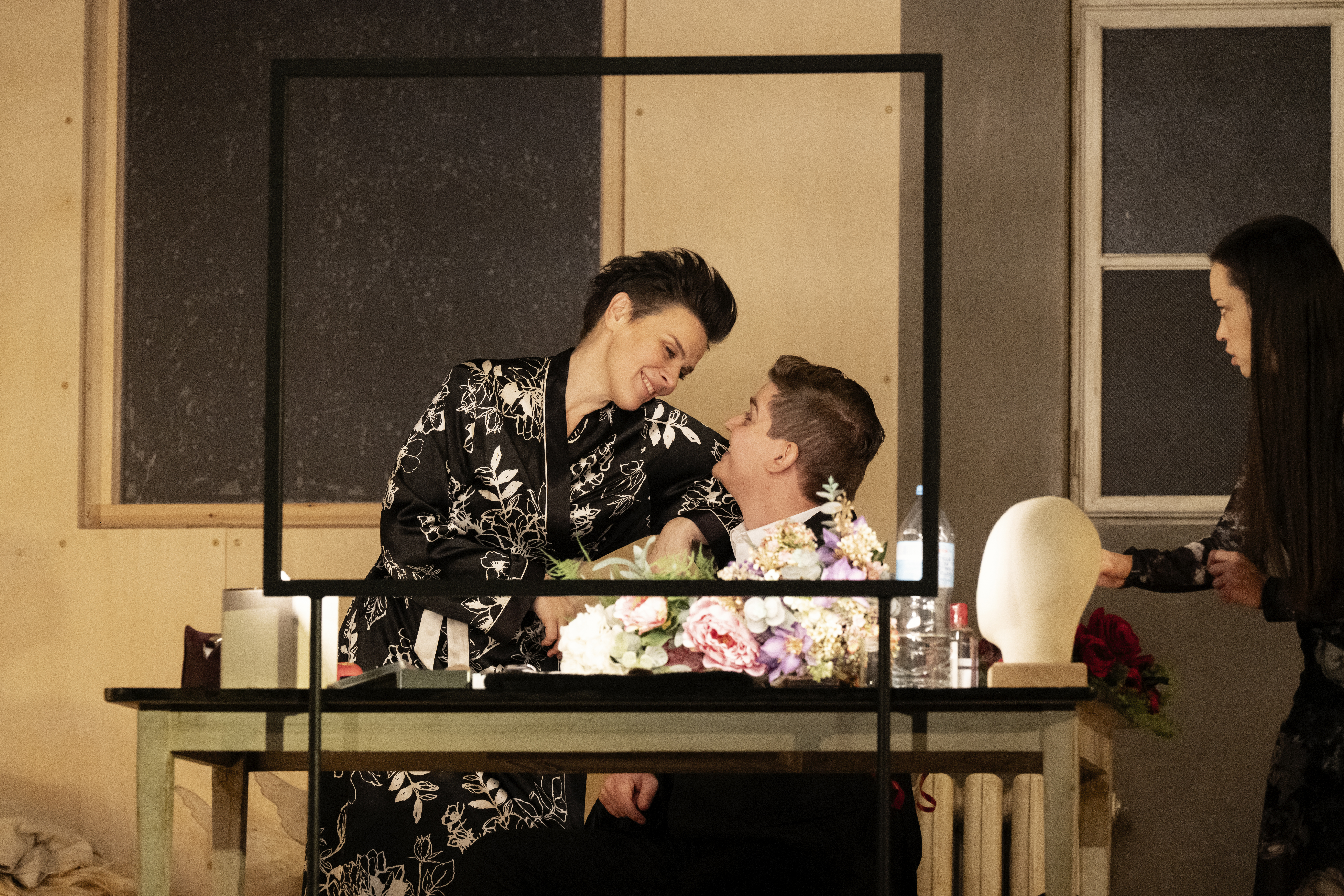 A man sits at a dressing table. A woman in a floral dressing gown sits in his lap. 