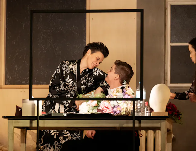 A man sits at a dressing table. A woman in a floral dressing gown sits in his lap.
