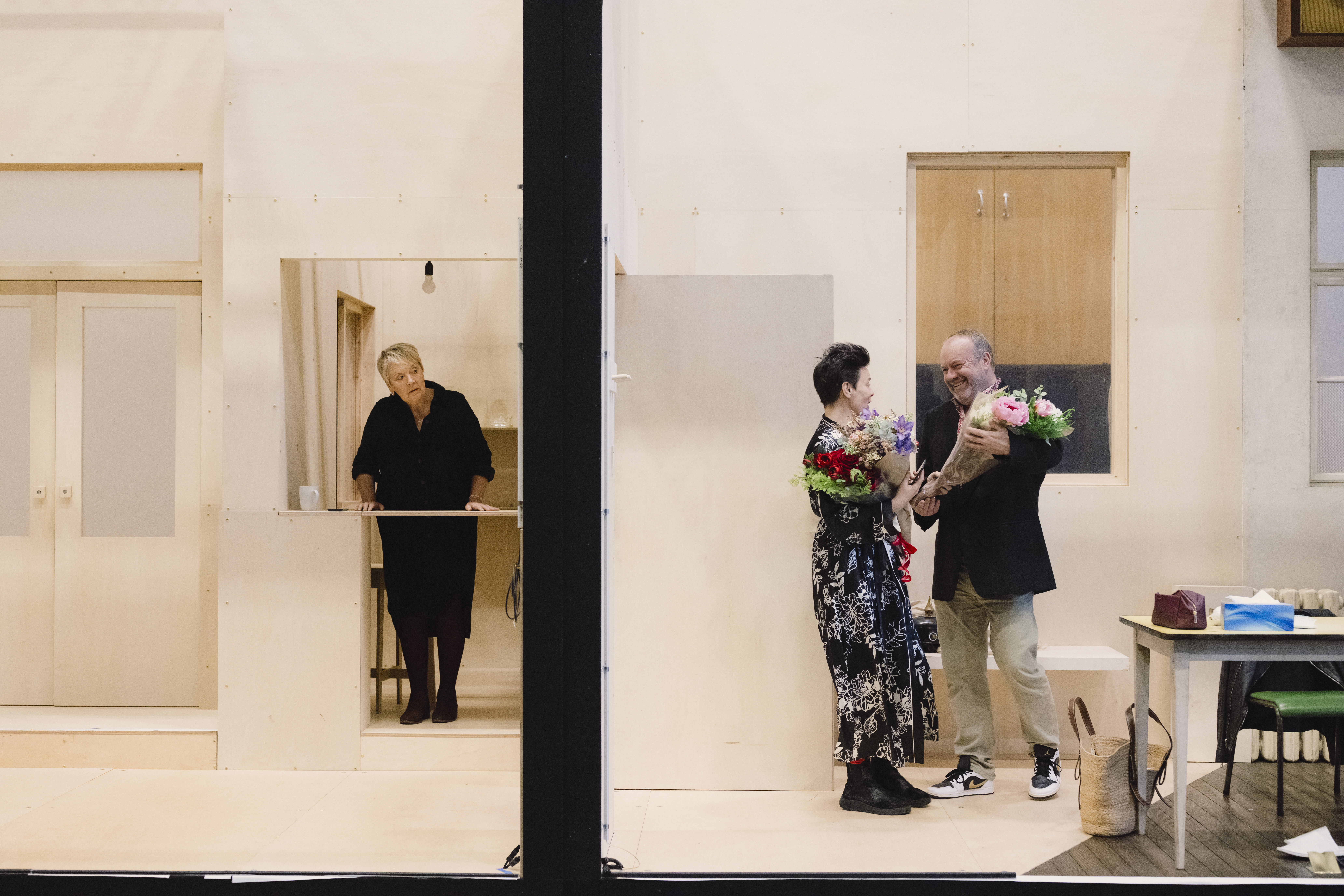 A man and a woman stand in a room. They are talking as both hold bouquets of flowers. The other side of the wall a woman in a black dress tried to listen in. 