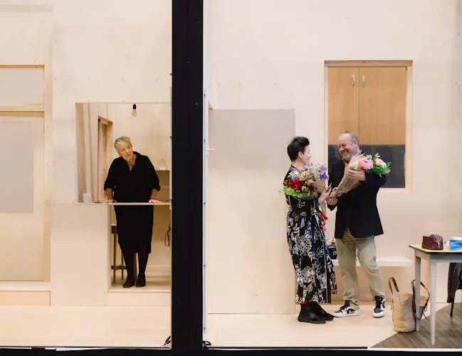 A man and a woman stand in a room. They are talking as both hold bouquets of flowers. The other side of the wall a woman in a black dress tried to listen in.