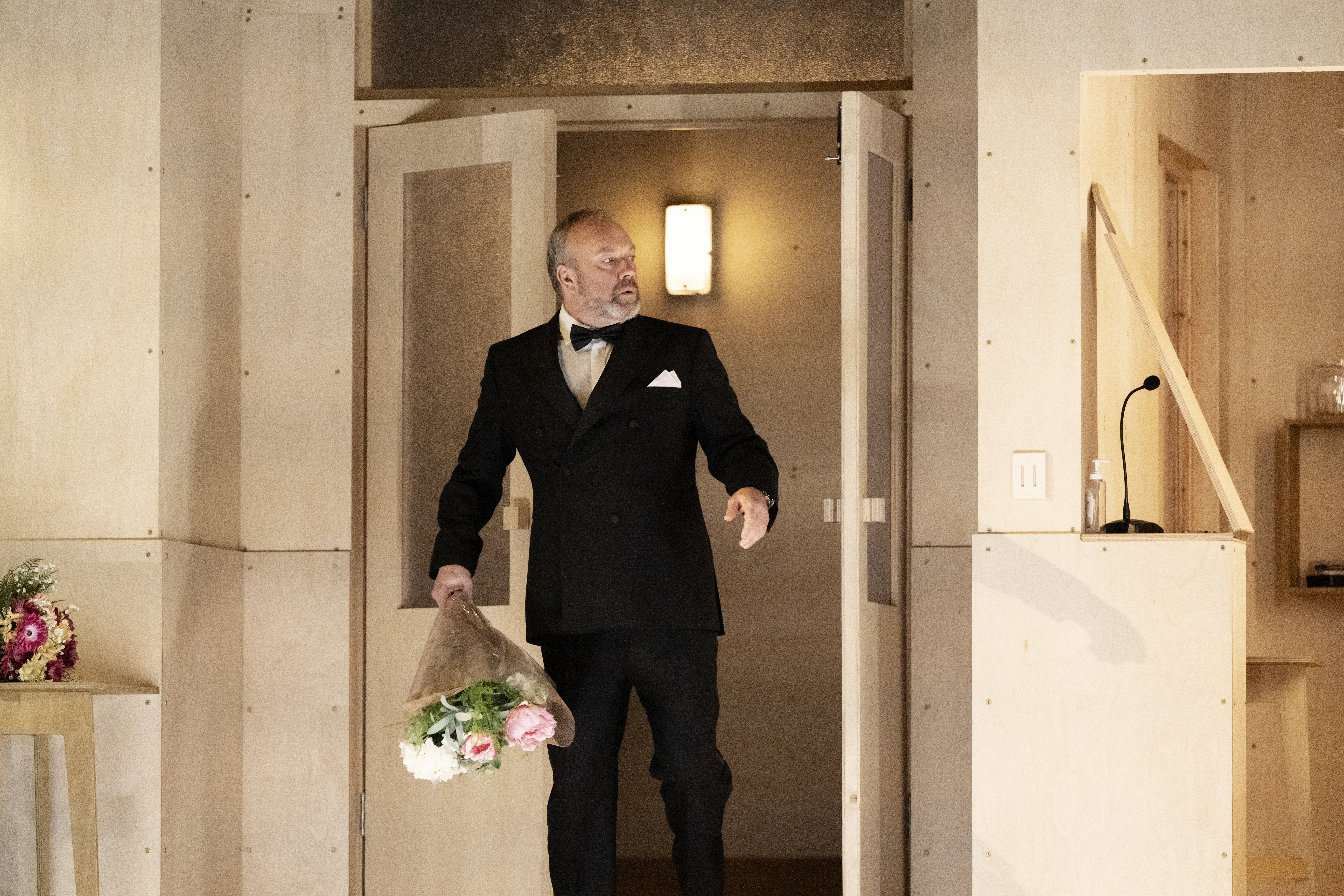 A man enters a room. He is wearing a black tuxedo and carring a bouquet of flowers. 