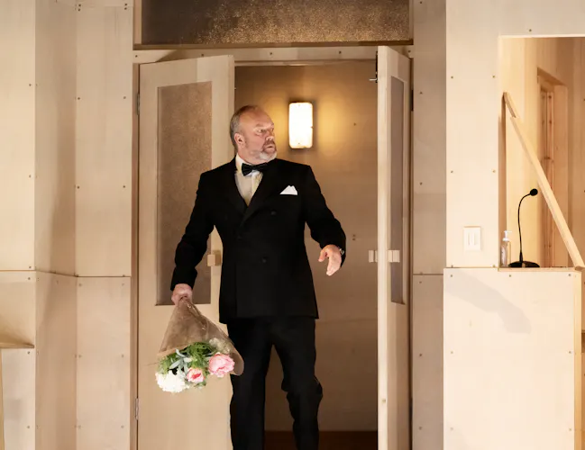 A man enters a room. He is wearing a black tuxedo and carring a bouquet of flowers.