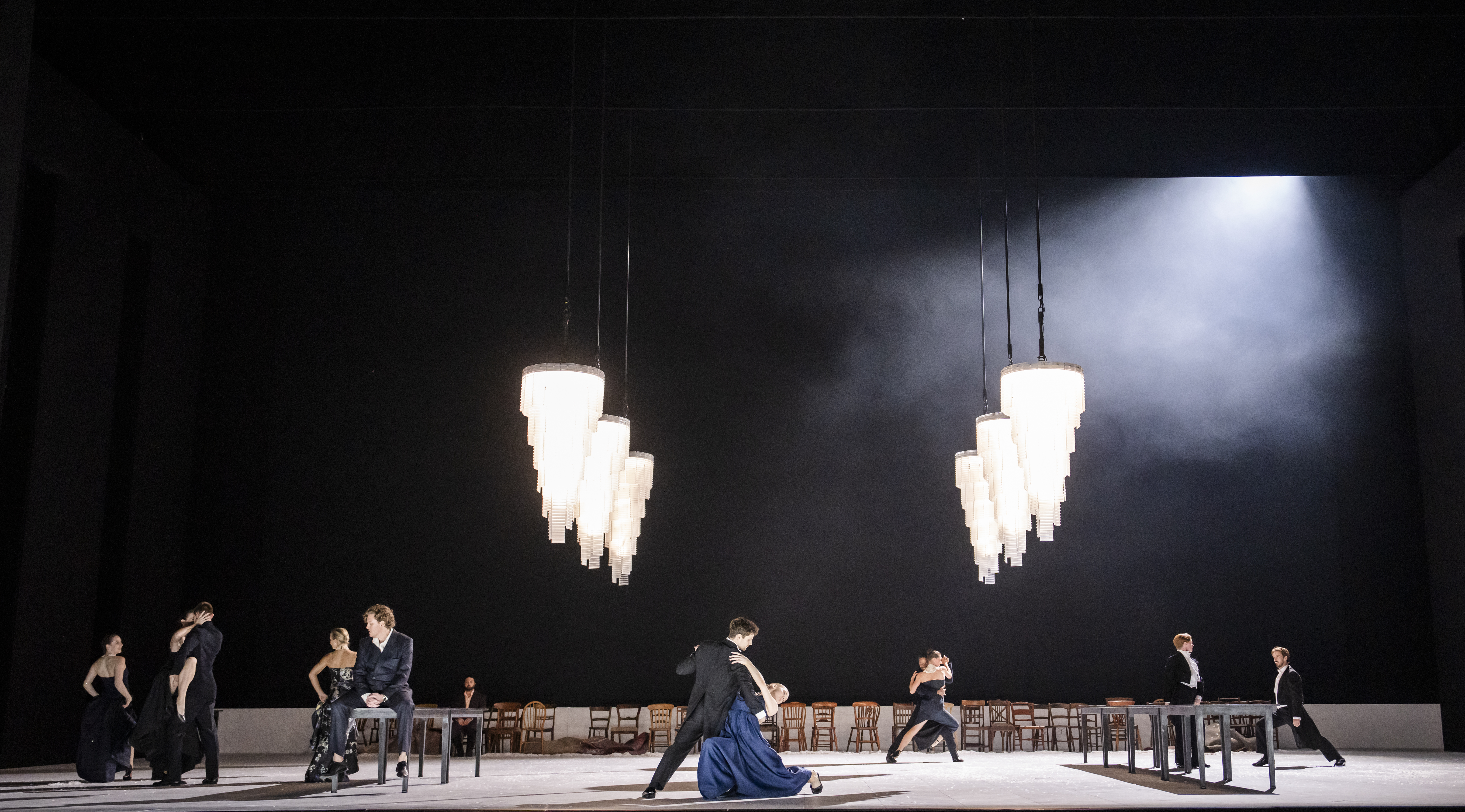 A couple ballroom dances on a stage. They are posed centrestage as the man wearing a black tuxedo suit dips the woman who is wearing a blue dress and white silk gloves. There are large crystal chandeliers hanging over them. Wooden chairs line the back of the stage.