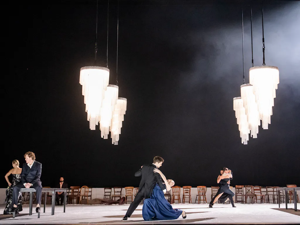 A couple ballroom dances on a stage. They are posed centrestage as the man wearing a black tuxedo suit dips the woman who is wearing a blue dress and white silk gloves. There are large crystal chandeliers hanging over them. Wooden chairs line the back of the stage.