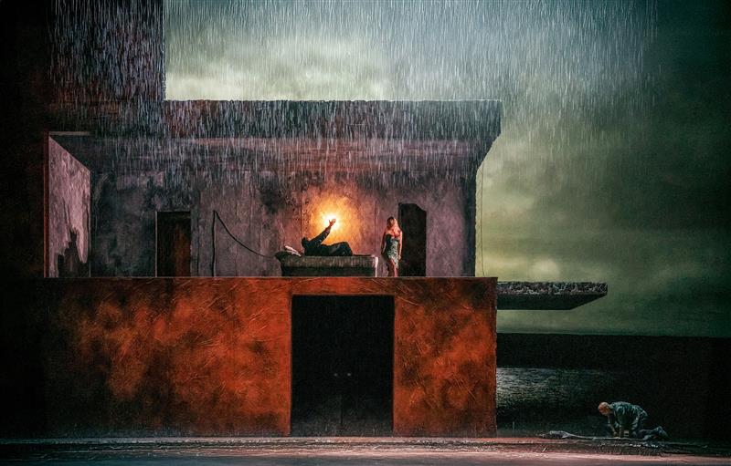 A darkened stage with rain water falling from above. In the centre is a two story building, on the upper floor a man lies in a bed with a light behind him, a woman stands over him. Outside the house a man kneels over a body lying on the floor. A stormy lake can be seen painted on the backdrop behind the house.