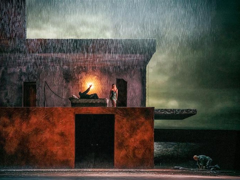 A darkened stage with rain water falling from above. In the centre is a two story building, on the upper floor a man lies in a bed with a light behind him, a woman stands over him. Outside the house a man kneels over a body lying on the floor. A stormy lake can be seen painted on the backdrop behind the house.