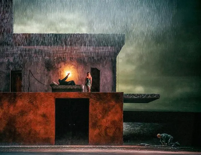 A darkened stage with rain water falling from above. In the centre is a two story building, on the upper floor a man lies in a bed with a light behind him, a woman stands over him. Outside the house a man kneels over a body lying on the floor. A stormy lake can be seen painted on the backdrop behind the house.