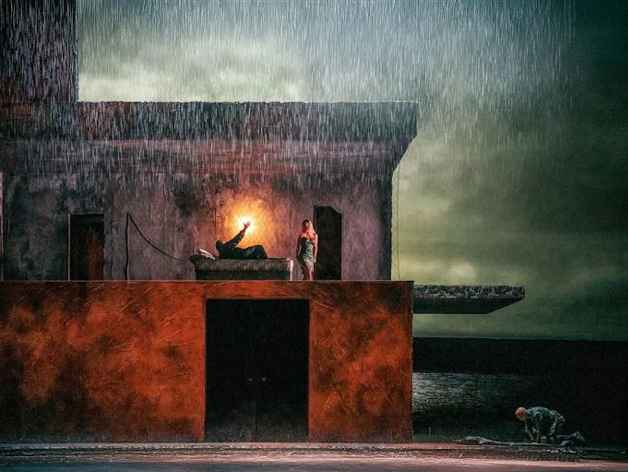 A darkened stage with rain water falling from above. In the centre is a two story building, on the upper floor a man lies in a bed with a light behind him, a woman stands over him. Outside the house a man kneels over a body lying on the floor. A stormy lake can be seen painted on the backdrop behind the house.