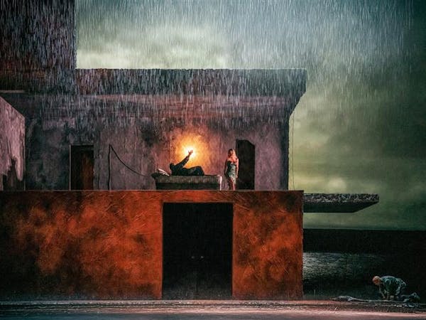 A darkened stage with rain water falling from above. In the centre is a two story building, on the upper floor a man lies in a bed with a light behind him, a woman stands over him. Outside the house a man kneels over a body lying on the floor. A stormy lake can be seen painted on the backdrop behind the house.