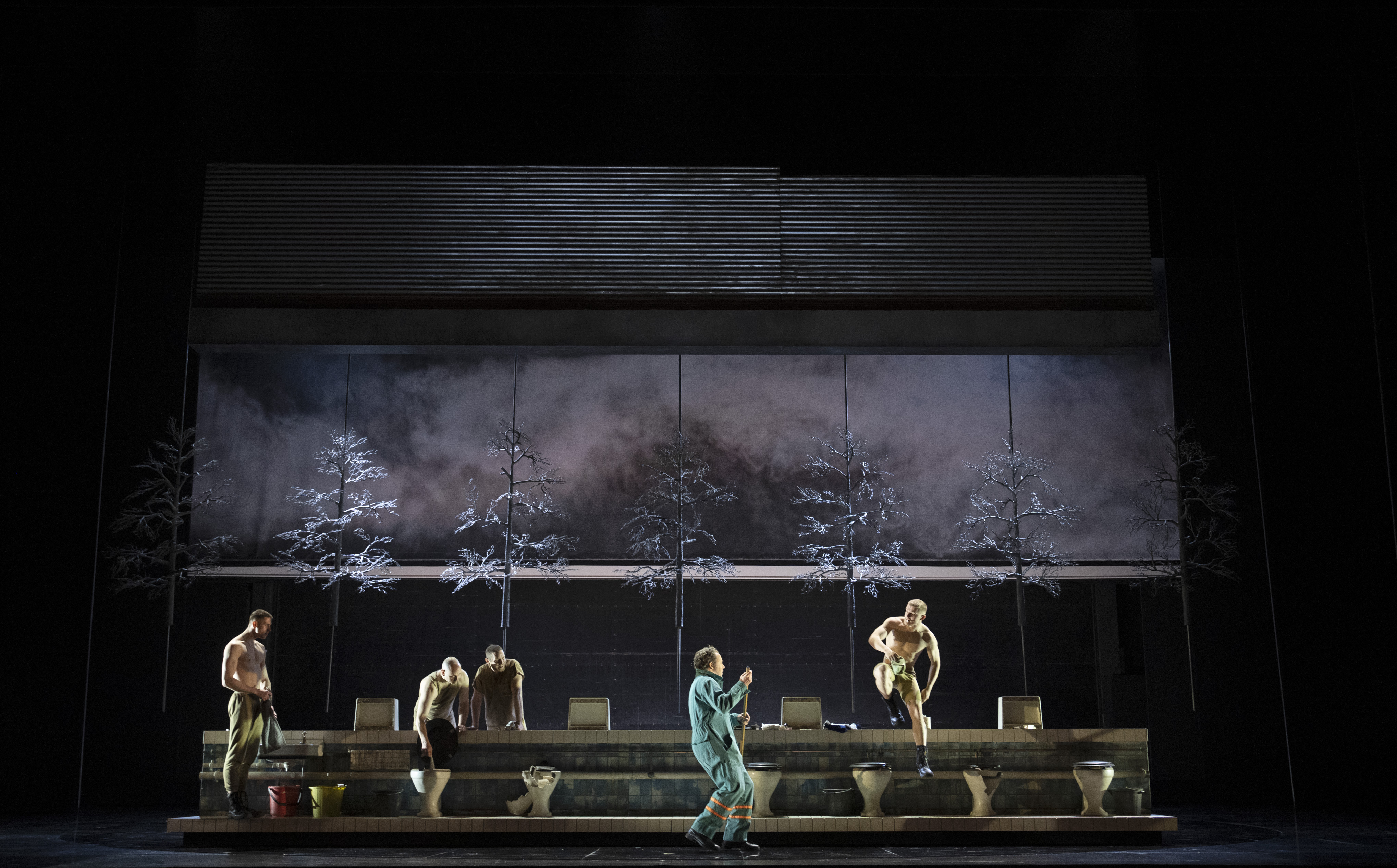 four shirtless performers in pants posing or sitting on a set with toilets, while a person in blue coveralls stands at center with a broom. The backdrop shows trees and a misty sky.