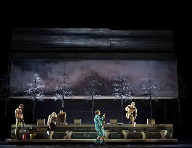 four shirtless performers in pants posing or sitting on a set with toilets, while a person in blue coveralls stands at center with a broom. The backdrop shows trees and a misty sky.