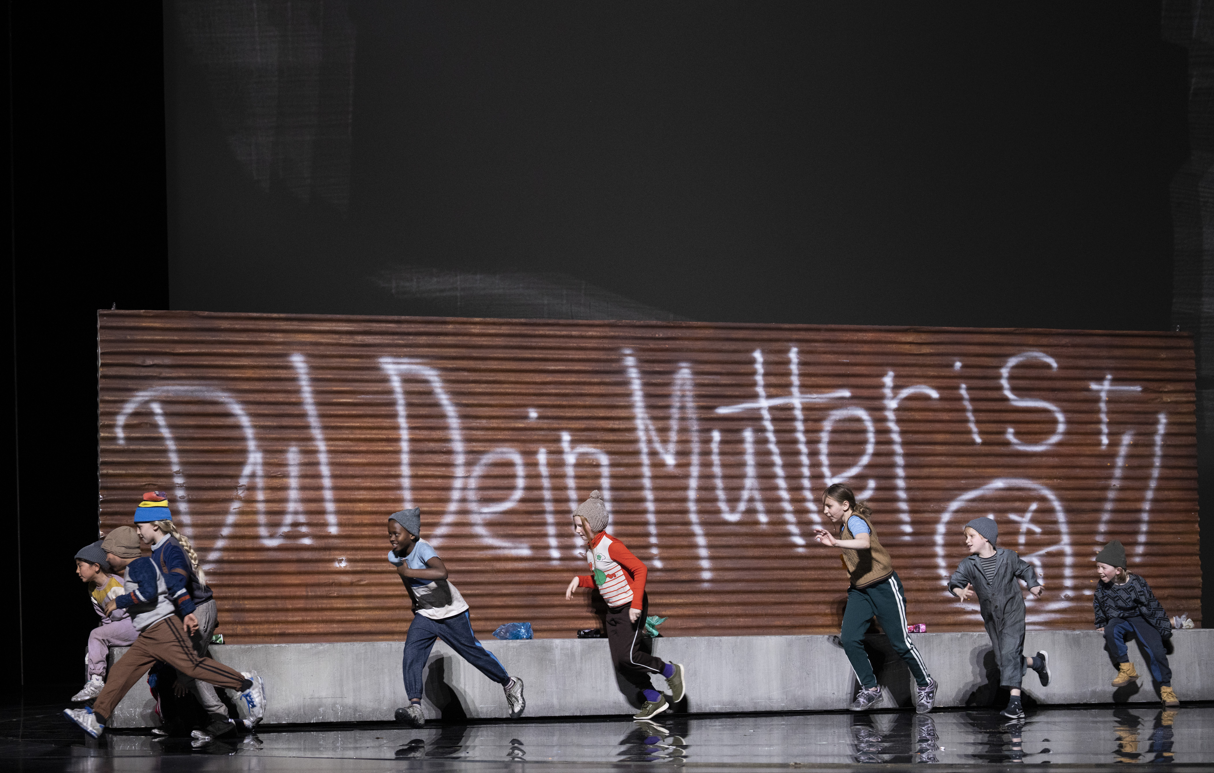 	 A group of children in winter clothes run past a wooden wall with large white graffiti that reads Du! Dein Mutter ist! The scene is set on a stage with a dark background.