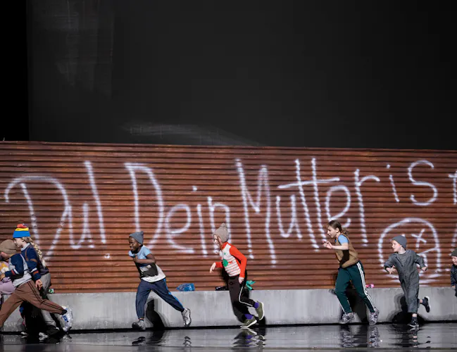 A group of children in winter clothes run past a wooden wall with large white graffiti that reads Du! Dein Mutter ist! The scene is set on a stage with a dark background.