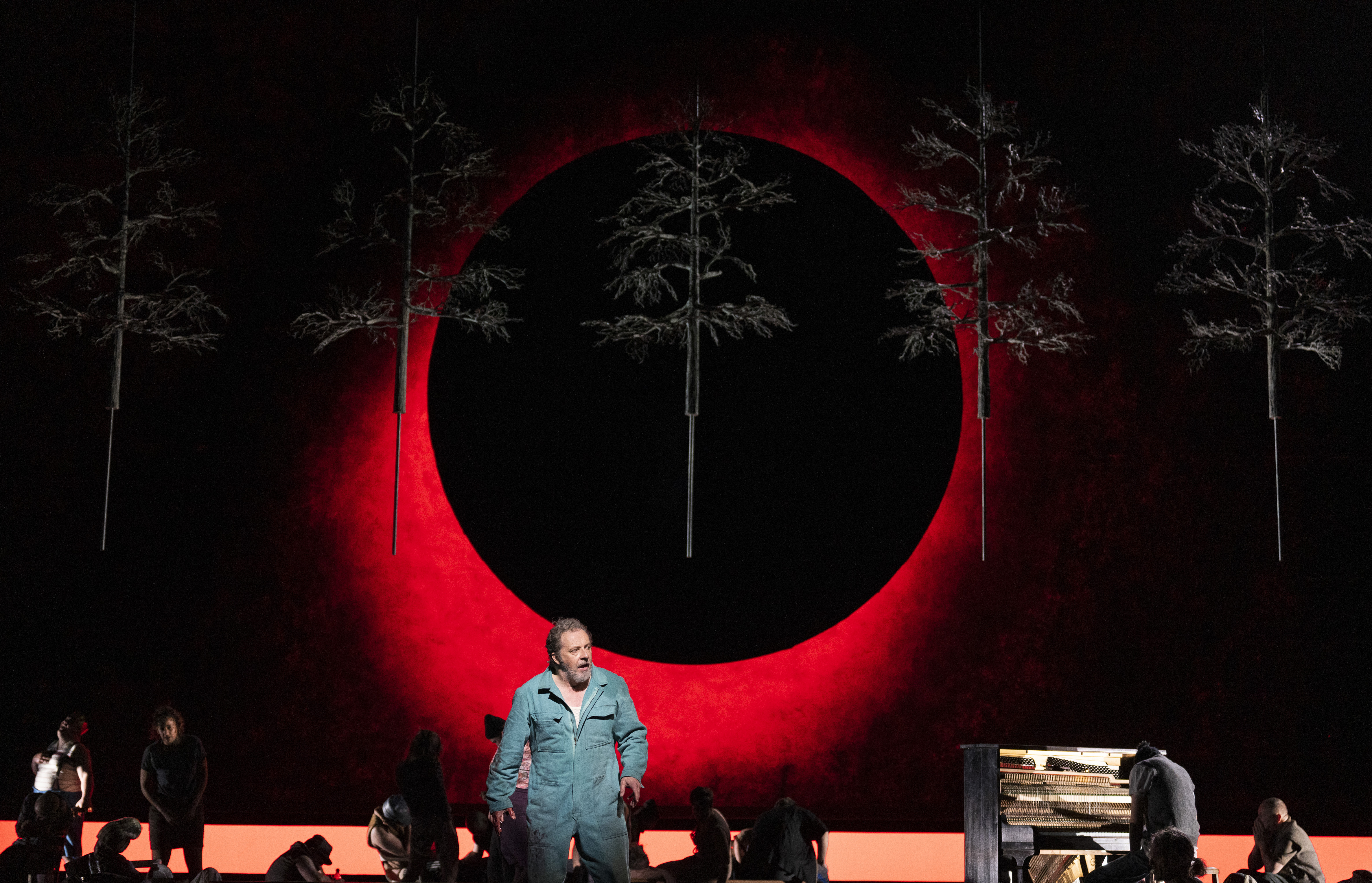 	 A man in a blue jumpsuit stands center stage in front of a large red and black circular backdrop, with suspended tree sculptures above and several people working or sitting on the stage around him.