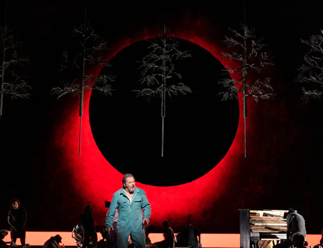 A man in a blue jumpsuit stands center stage in front of a large red and black circular backdrop, with suspended tree sculptures above and several people working or sitting on the stage around him.